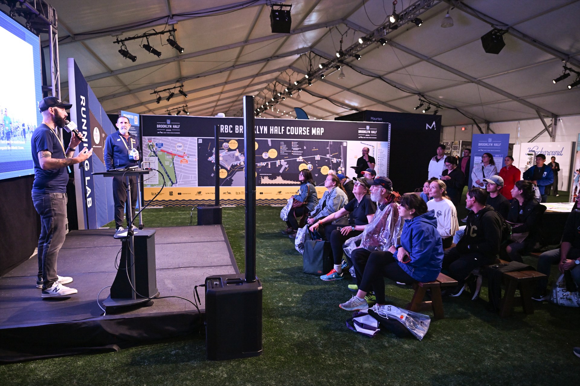 NYRR run coaches  on a stage speak to a group of people seated on benches at the Running Lab at the 2025 RBC Brooklyn Half Pre-Party