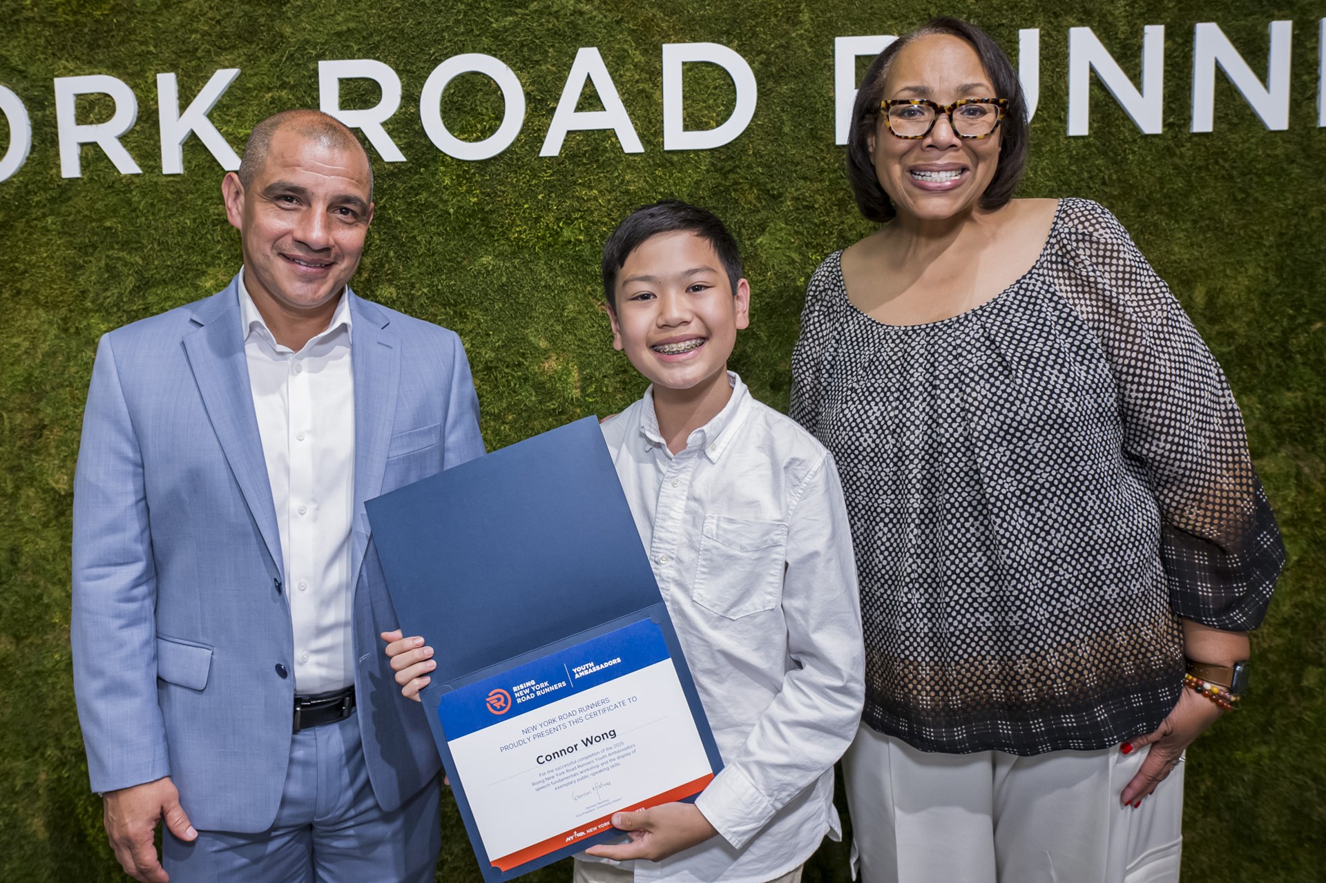 A youth ambassador smiles with his certificate and two NYRR employees at graduation