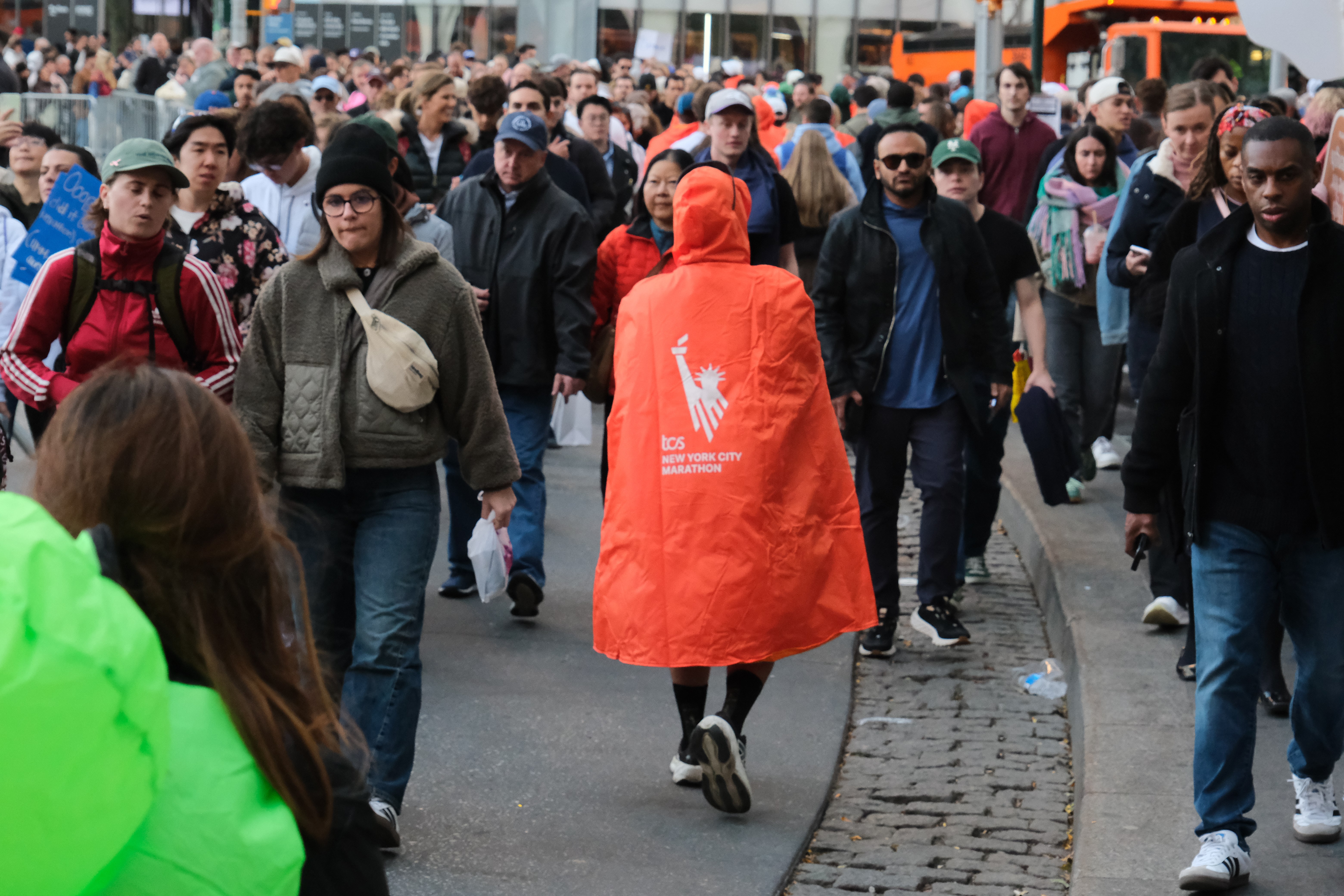 Marathon after 2025 TCS NYC Marathon wearing orange poncho facing away from camera on crowded street.